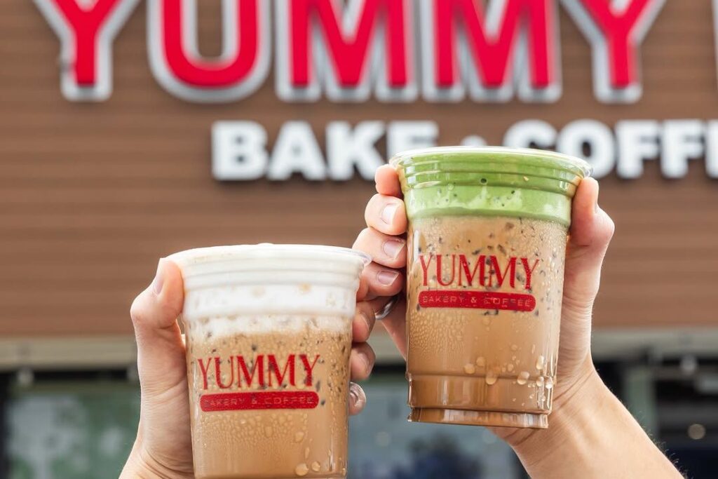 Two hands holding Yummy iced coffee drinks in front of the Yummy Bake and Coffee storefront sign