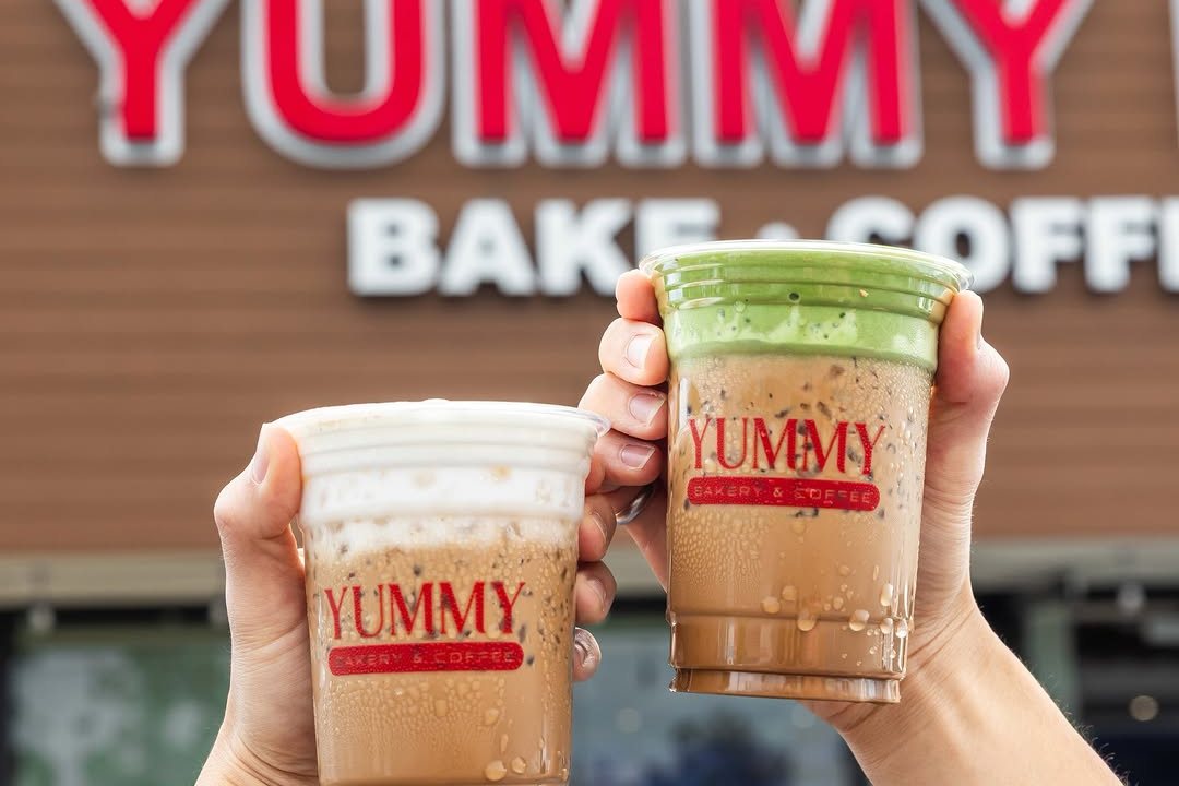 Two hands holding Yummy iced coffee drinks in front of the Yummy Bake and Coffee storefront sign
